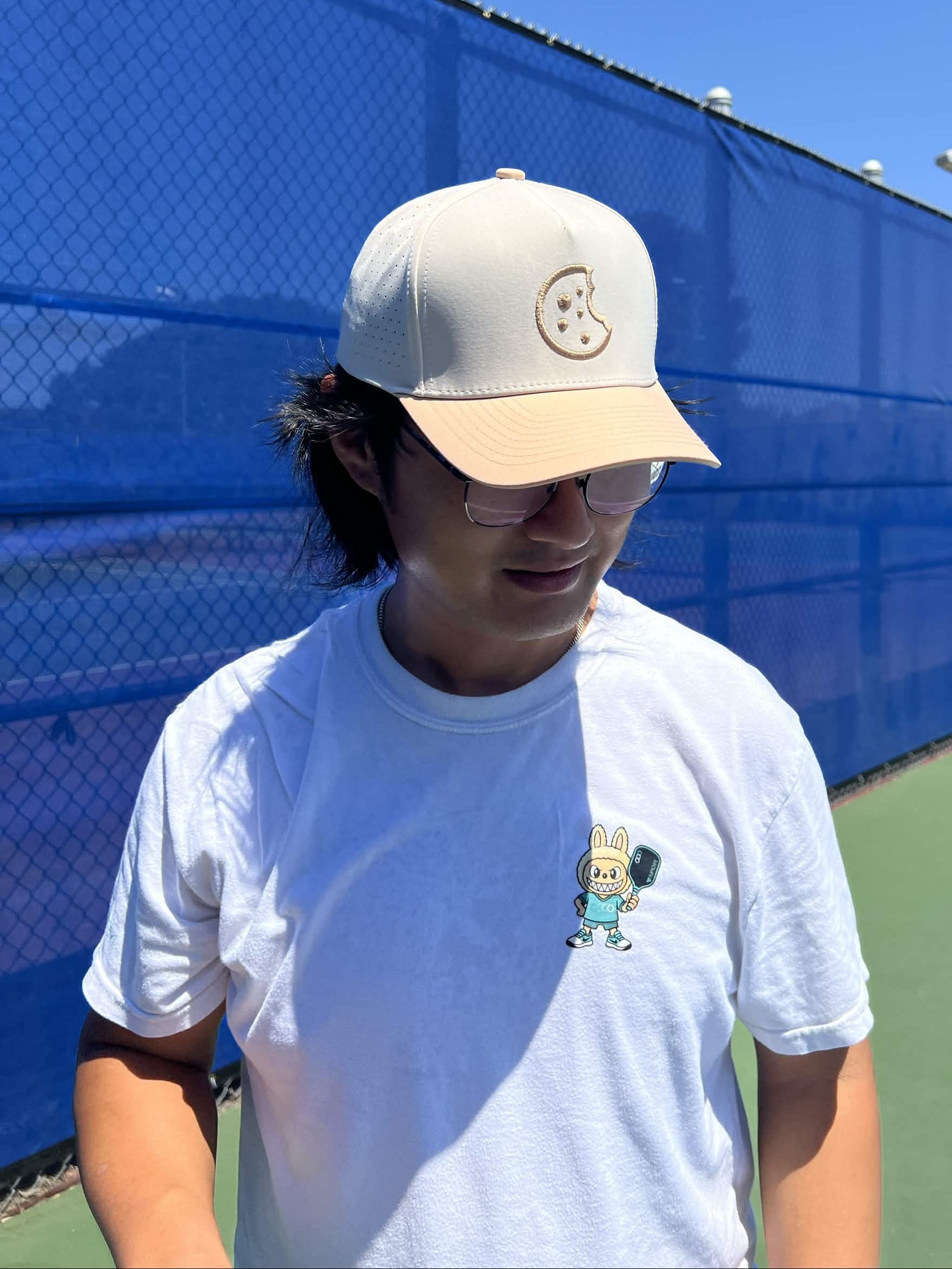 A man wearing a white t-shirt with a small cartoon bunny graphic on the chest, a beige baseball cap, and glasses, standing on an outdoor tennis court with a blue fence in the background.