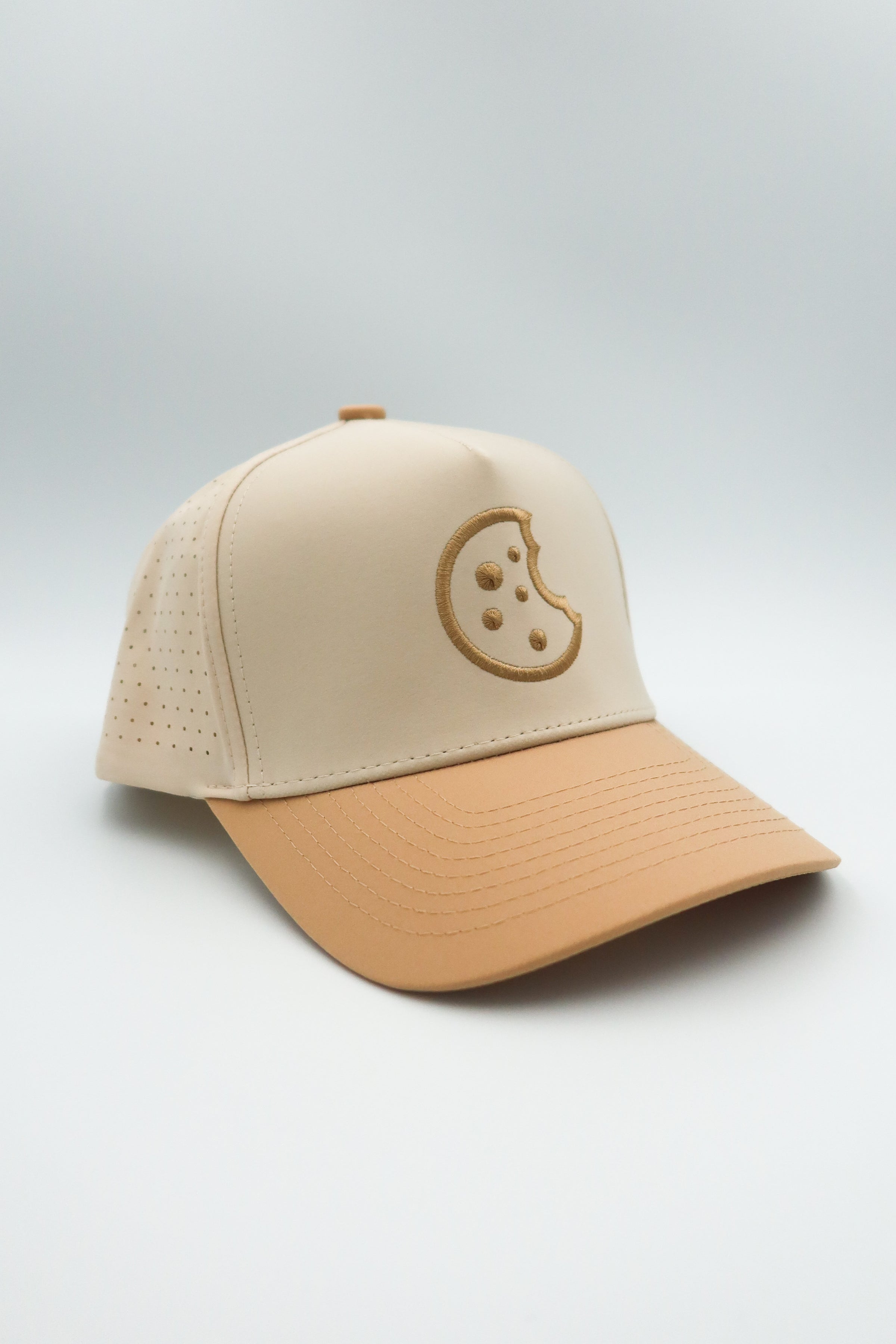 A structured baseball cap with a light beige crown and a tan visor, featuring a tonal embroidered cookie graphic on the front, set against a neutral light gray background.