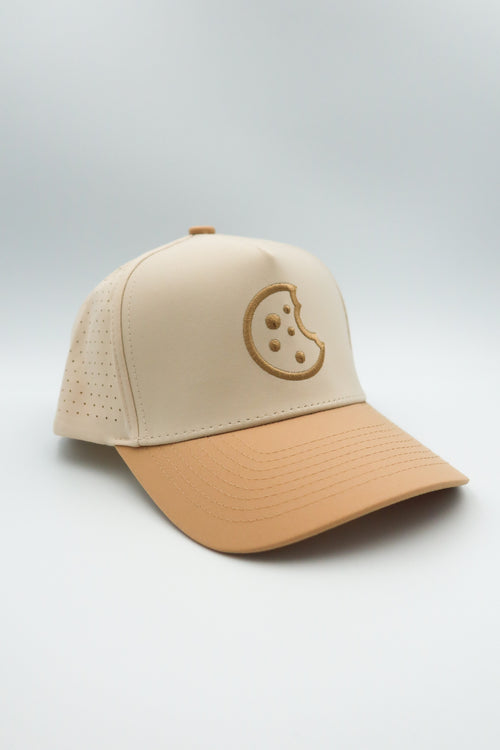 A structured baseball cap with a light beige crown and a tan visor, featuring a tonal embroidered cookie graphic on the front, set against a neutral light gray background.