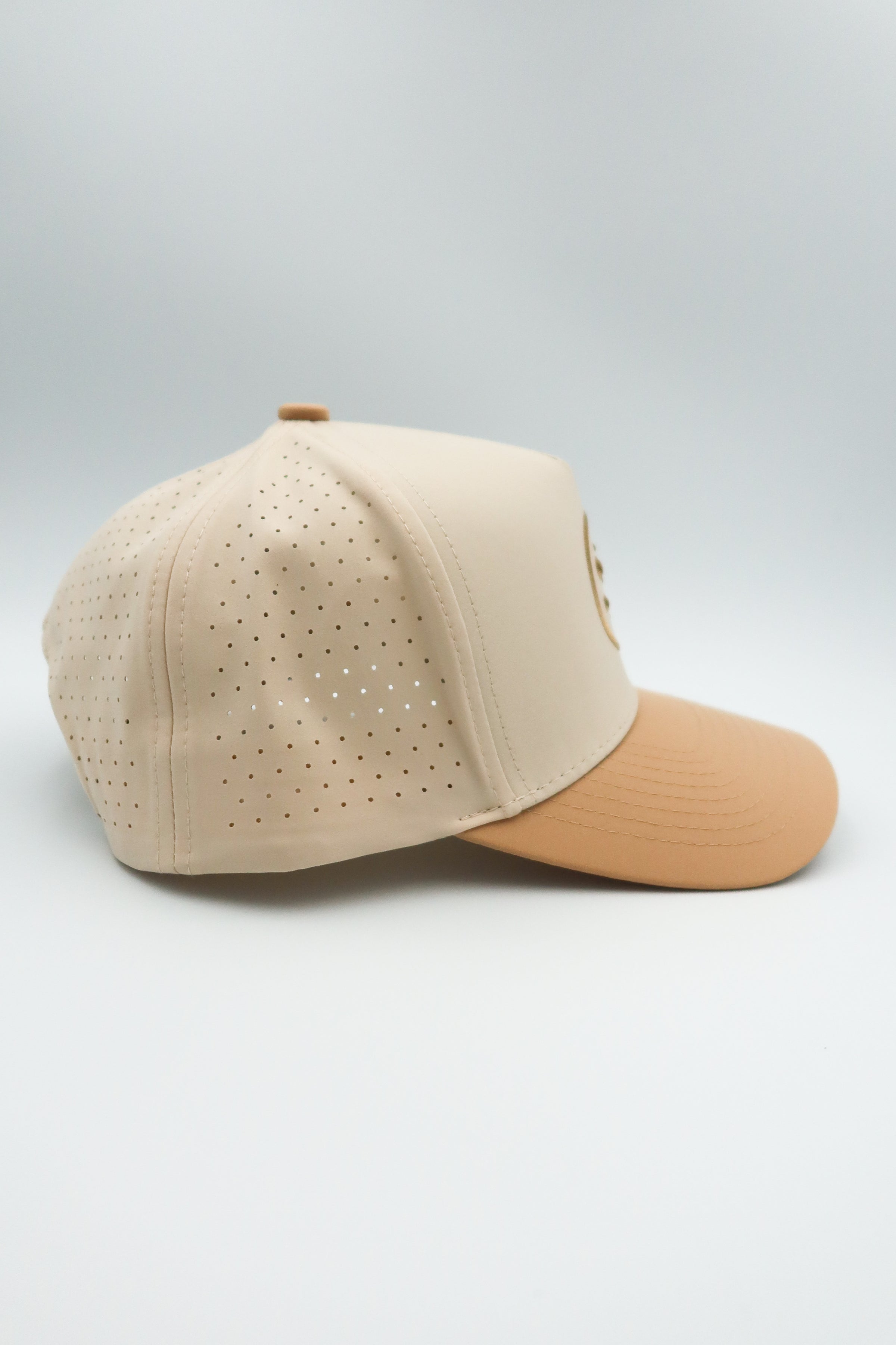 A structured baseball cap in light beige with perforated side panels and a contrasting tan visor, shown on a light gray studio background.