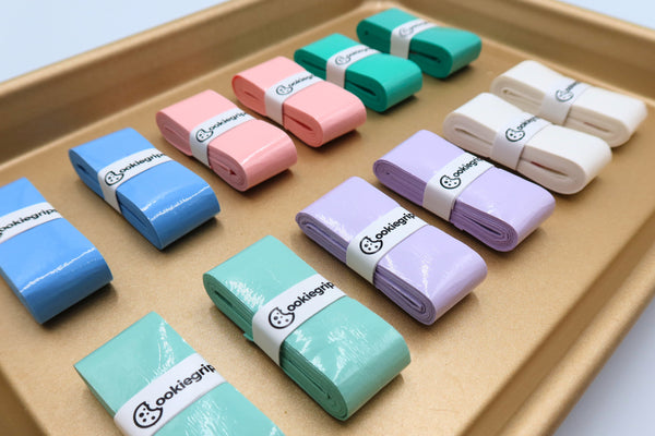Assorted pastel cookiegrips pickleball overgrips displayed on a baking tray — colors include pink, lavender, mint, and teal, each wrapped with a cookiegrips logo band.